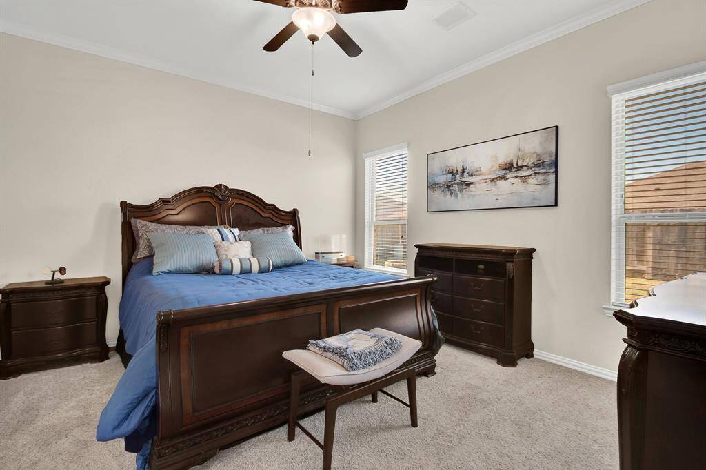 This bedroom features a spacious layout with a large bed, elegant wooden furniture, and ample natural light from two windows. Neutral walls and a ceiling fan add to the comfortable atmosphere.
