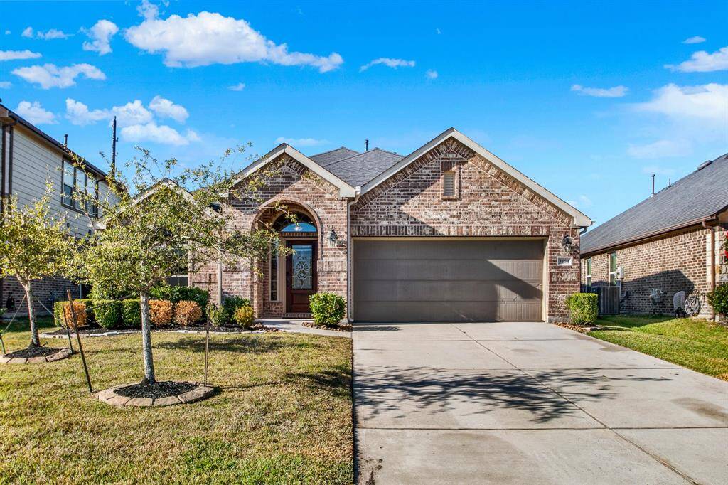 This is a charming brick single-story home with a two-car garage and a well-maintained front yard, featuring a mature tree and landscaped bushes. The entrance is inviting with an arched doorway and a decorative glass front door.