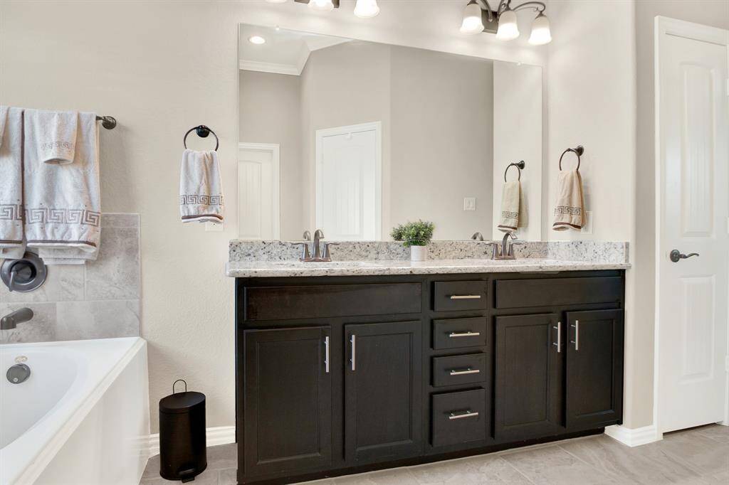 This bathroom features a double vanity with a granite countertop and dark wood cabinets. It includes a large mirror, stylish lighting, and a bathtub with tiled accents. The space is bright and modern, offering a clean and elegant design.