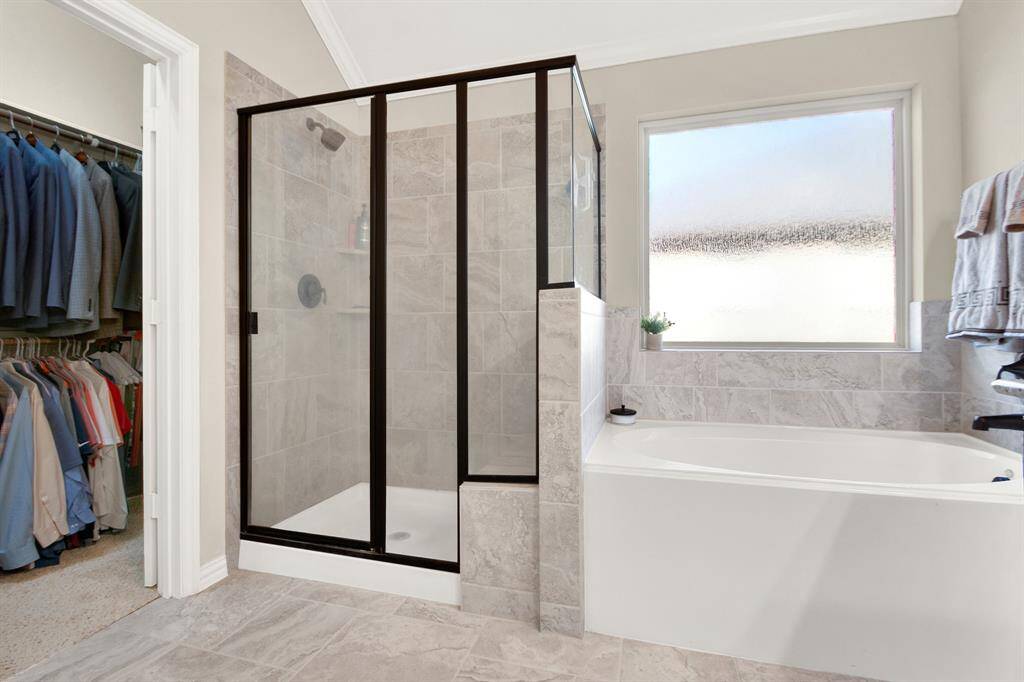 This photo shows a modern bathroom with a spacious glass-enclosed shower and a separate bathtub. It features elegant tile work and a frosted window, with an adjoining walk-in closet visible to the side.