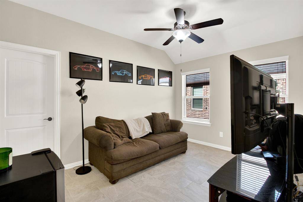 This cozy living room features a comfortable brown sofa, modern ceiling fan, and large TV. It has decorative car-themed wall art and ample natural light from two windows, creating a bright and inviting space.