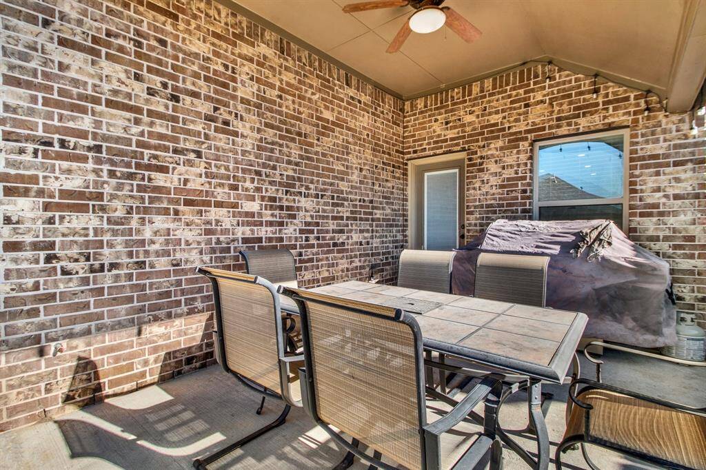 This photo shows a cozy outdoor patio with a brick wall, featuring a ceiling fan and a covered grill. There's a dining table with six chairs, perfect for outdoor meals and entertaining.