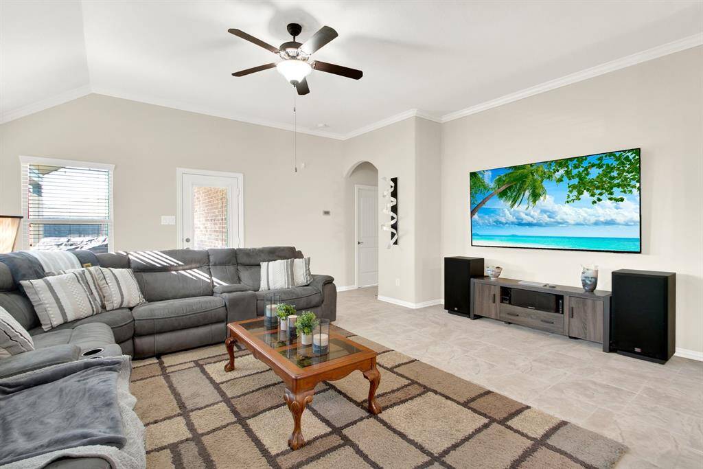This living room is spacious and bright, featuring a large sectional couch, a wall-mounted TV, and a ceiling fan. The neutral walls and tile flooring create a cozy and welcoming atmosphere, ideal for relaxation and entertainment.