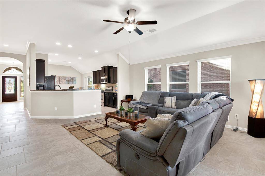 This spacious living area features an open floor plan with a modern kitchen, ample natural light from large windows, and a cozy sectional sofa. The neutral tile flooring and ceiling fan add comfort and style.