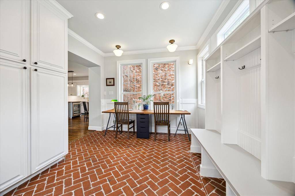 An additional view of the mudroom reveals a charming built-in workstation framed by tall windows, abundant cabinetry, and classic brick herringbone floors that extend seamlessly toward the kitchen.