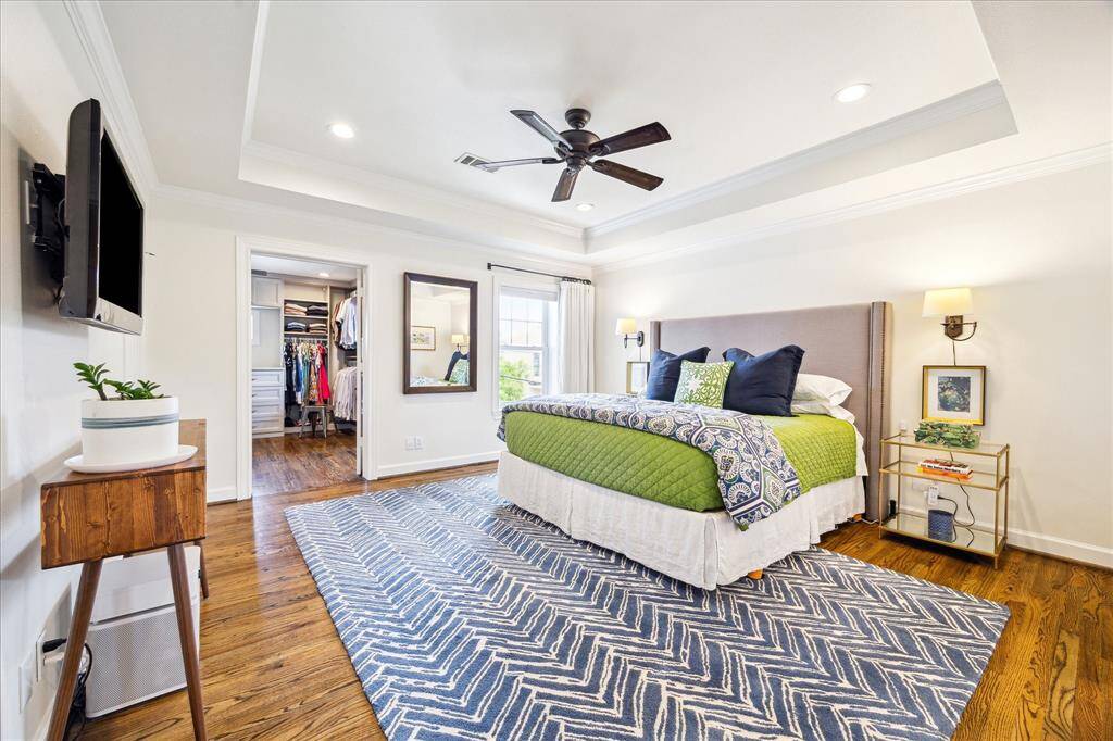 This elegant primary suite impresses with a tray ceiling, warm wood floors, abundant natural light, and a seamless flow into the custom walk-in closet and luxurious en-suite bath.