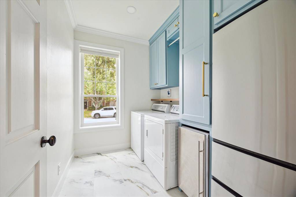 This additional view of the laundry room showcases elegant marble-look tile flooring, custom sky-blue cabinetry with brass hardware, and a large window that fills the space with natural light.