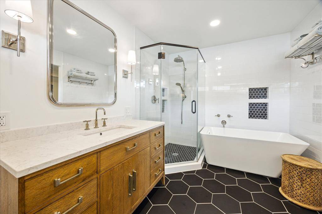 The spa-inspired primary bath exudes modern elegance with its freestanding soaking tub, frameless glass shower, striking hexagonal tile flooring, and rich wood vanity topped with sleek marble.