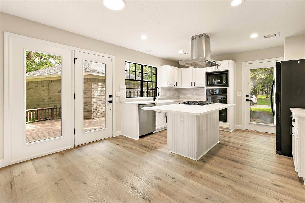 This modern kitchen features white cabinets, an island cooktop under a stainless steel hood, and light wood-look floors. Multiple glass doors open to outdoor spaces, enhancing the room's bright and airy atmosphere.
