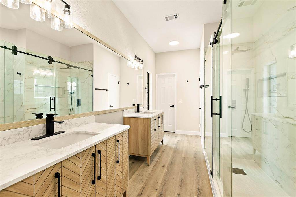 Luxurious primary bath with double vanity featuring chic chevron wood cabinetry, quartz counters, and sleek black fixtures. Expansive walk-in shower boasts large format marble-look tile and modern barn-style glass doors. Bright and airy with abundant natural light