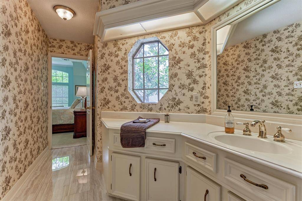 Private ensuite bath with charming floral wallpaper, abundant cabinet space, and a unique octagonal window for added character.