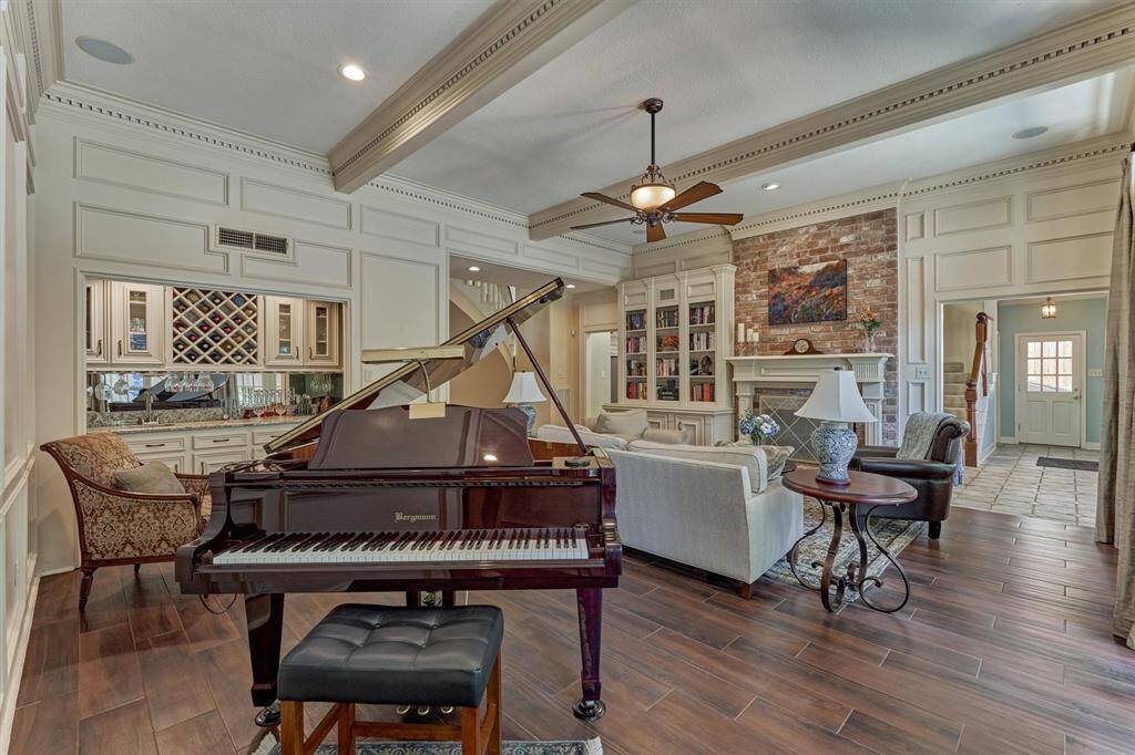 Elegant living space featuring rich wood-look tile, grand piano area, custom built-ins, brick fireplace, and a stunning wet bar with wine storage.