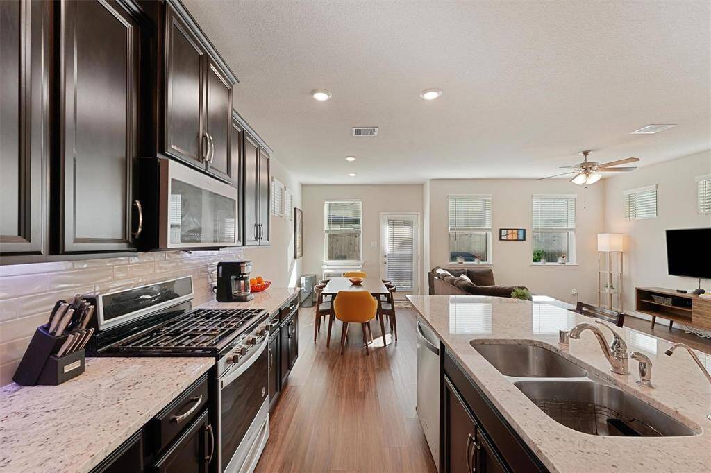 Modern, open-concept kitchen and living area. The kitchen has dark cabinetry, granite countertops, and stainless steel appliances. It flows seamlessly into a bright living space with large windows, wood flooring, and a ceiling fan, creating a cozy and inviting atmosphere.