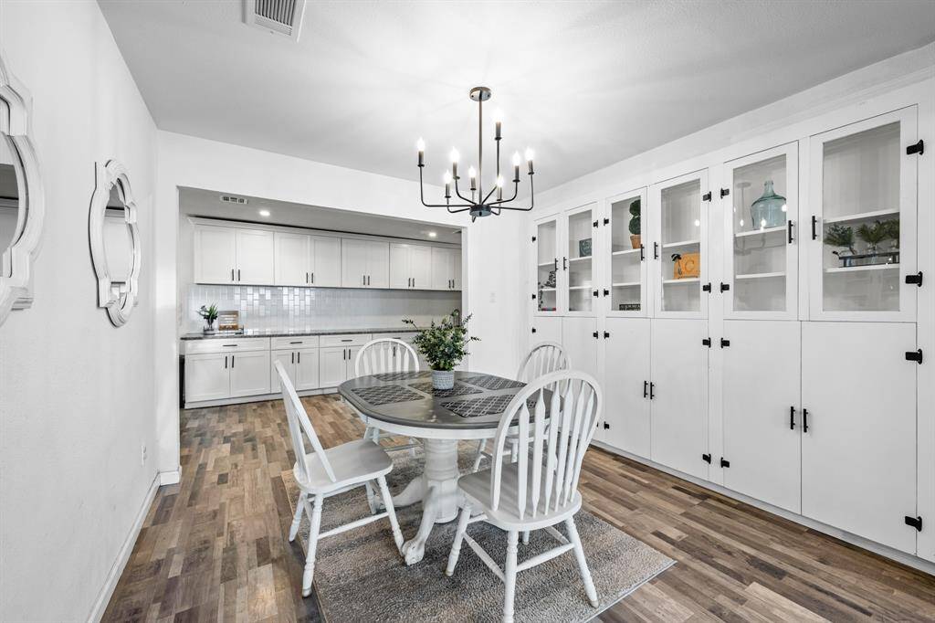 This bright dining area features vinyl plank floors, built-in cabinetry with glass door fronts, and a modern chandelier. The open layout connects seamlessly to a sleek kitchen, offering ample storage and display space.