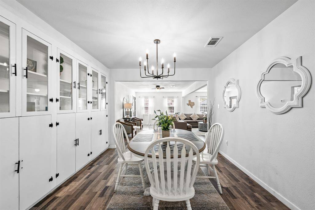 This photo showcases a bright dining area with built-in cabinets, a modern chandelier, and a round table with chairs. The space opens into a cozy living room, featuring wood-look flooring and decorative wall accents.