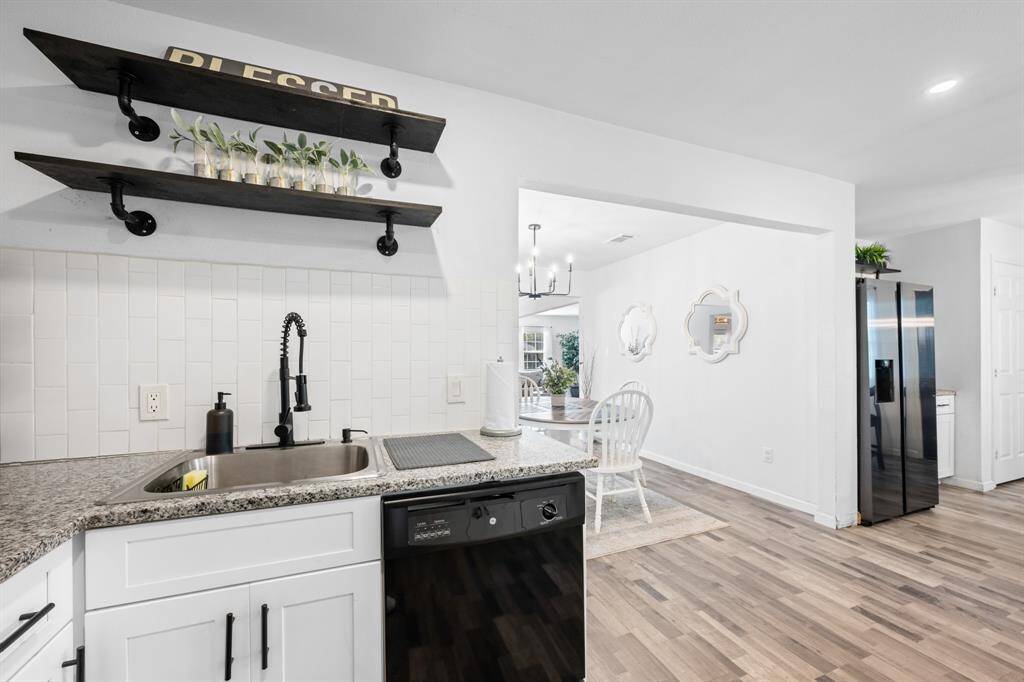 This photo showcases a modern kitchen with white cabinetry and a granite countertop, featuring a sleek black faucet. Open shelving with decorative items adds charm, while an adjoining dining area with natural light enhances the inviting atmosphere.