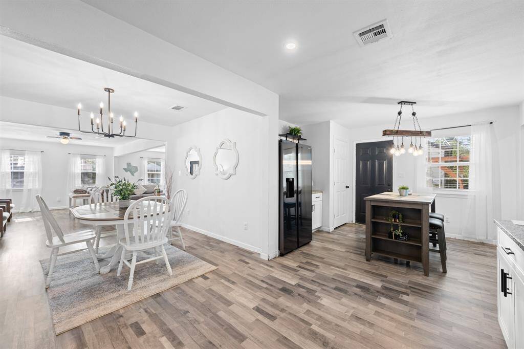 This photo showcases a bright, open-concept dining and kitchen area featuring modern lighting, white walls, and wood-look flooring. The dining area has a round table with white chairs, while the kitchen includes a central island and large windows for natural light.