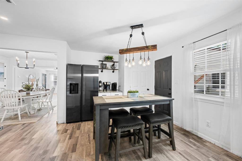 This photo showcases a modern kitchen with a sleek black refrigerator, a breakfast table with seating, and stylish lighting. The space is open and bright, with a dining area visible in the background, featuring a chandelier and a wooden table set.