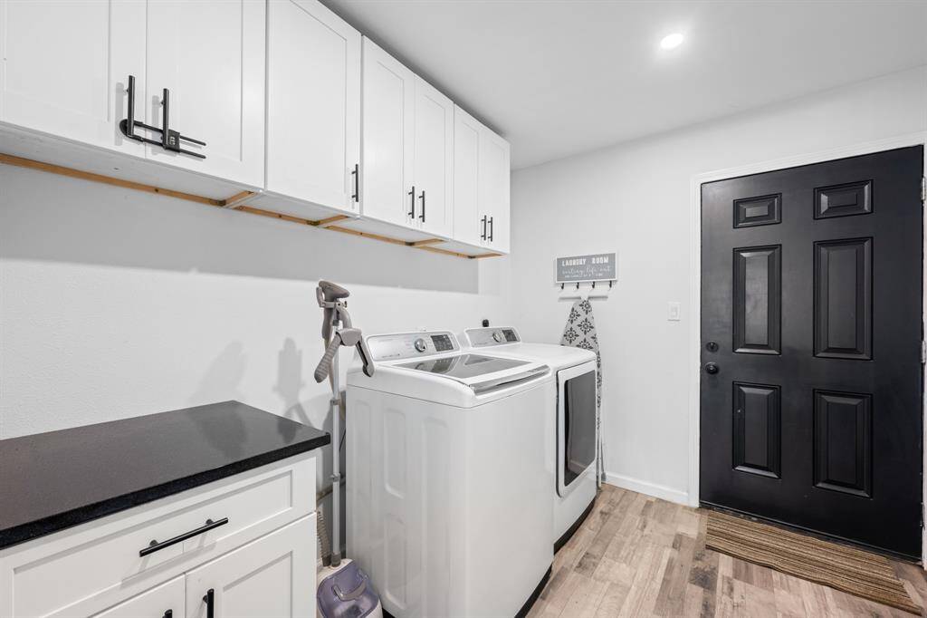 This photo shows a modern laundry room featuring a washer and dryer set. The space has white cabinetry with black handles, a black countertop, and a black door. The room is well-lit and has wood-style flooring, creating a clean and functional environment.
