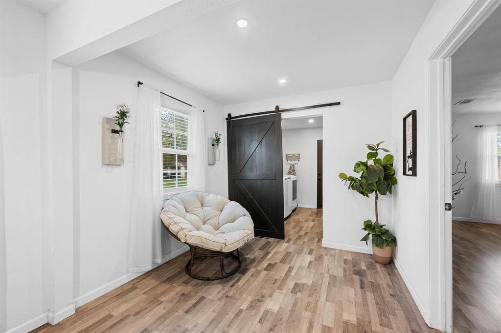 This bright, inviting space features hardwood-look floors, a cozy chair by a window with sheer curtains, a stylish barn door, and tasteful decor including plants, creating a warm and welcoming atmosphere.