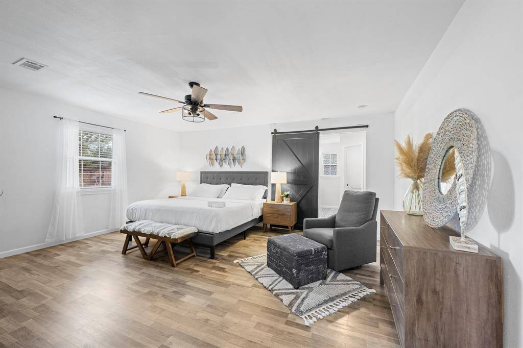 This spacious bedroom features a modern design with a cozy atmosphere. It includes a comfortable bed, stylish furniture, and a sliding barn door leading to an adjacent area. Natural light fills the room through a large window, highlighting the sleek vinyl plank flooring.