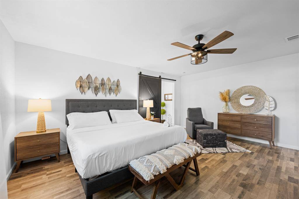 This inviting bedroom features a spacious layout with modern decor, a comfortable bed, stylish wooden furniture, and a ceiling fan. Natural light enhances the cozy, serene atmosphere, perfect for relaxation.