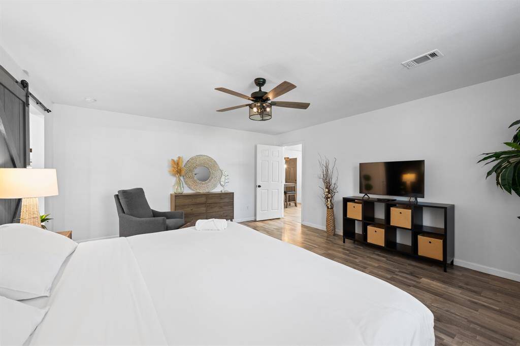 This spacious bedroom features modern decor with a ceiling fan, ample natural light, and a neutral color palette. It includes a cozy seating area, TV stand, and stylish storage, creating a comfortable and inviting atmosphere.