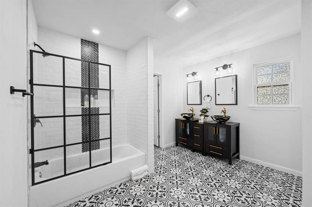 This bathroom features a modern design with a black-and-white color scheme. It includes a sleek shower with a glass partition, a double vanity with vessel sinks, and stylish patterned floor tiles. Ample natural light enters through a window, enhancing the bright and airy feel.
