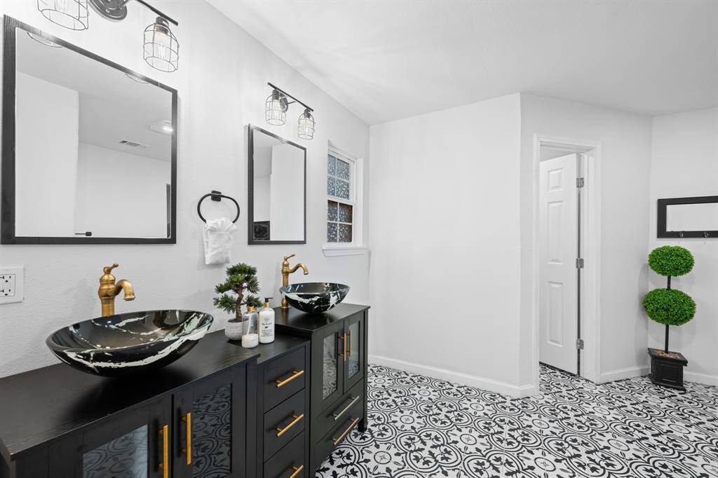 This modern bathroom features a stylish double vanity with vessel sinks, brass fixtures, and elegant black cabinetry. The floor showcases intricate black-and-white patterned tiles, and the room is well-lit with contemporary lighting.