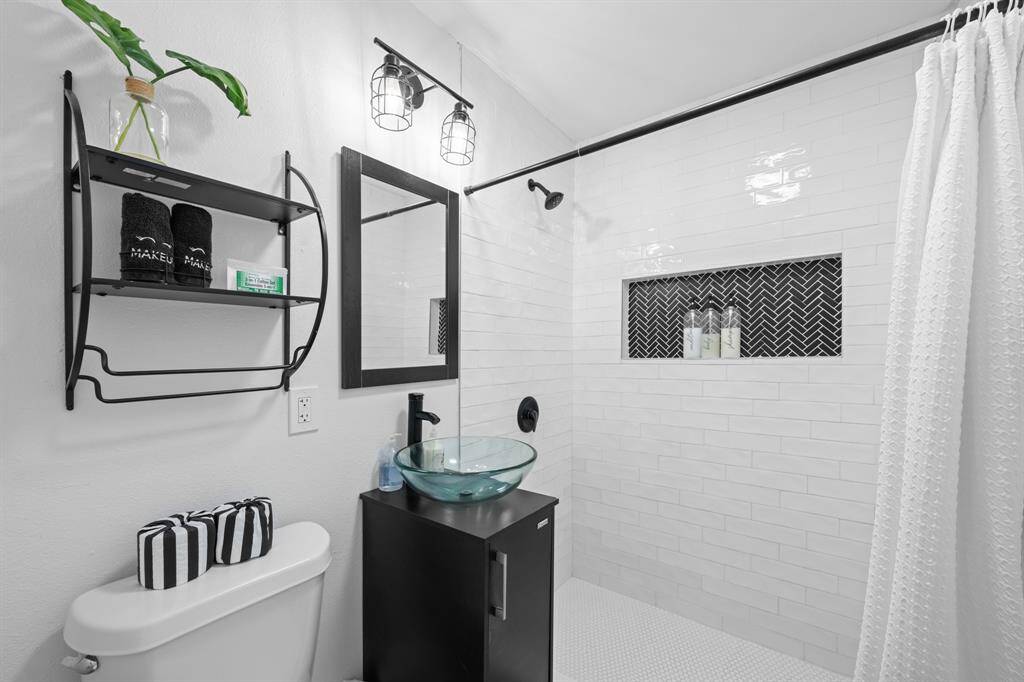 This bathroom features a modern design with sleek white tiles, a glass vessel sink, and a black vanity. There's a stylish shower area with a herringbone accent and a functional shelving unit. The space is bright and contemporary.