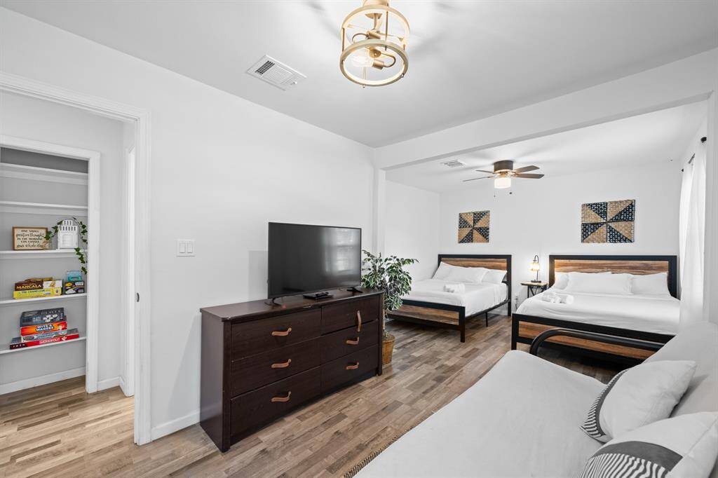 This spacious bedroom features two large beds, a cozy sitting area, and a modern ceiling fan. The room is bright with natural light, has wood-look flooring, and includes a TV and dresser. A built-in shelf provides extra storage space.