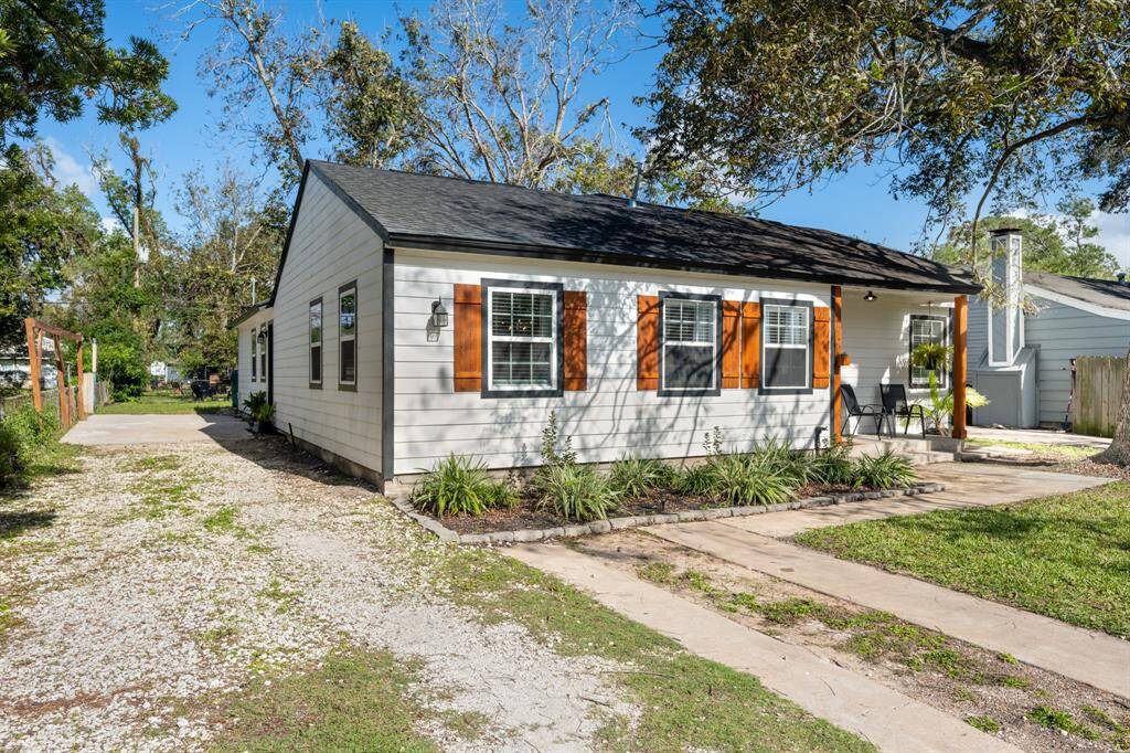 This charming single-story home features a cozy front porch, white siding with warm wood shutters, and a tidy landscape. A gravel driveway leads to the backyard, surrounded by mature trees, offering a serene and inviting atmosphere.