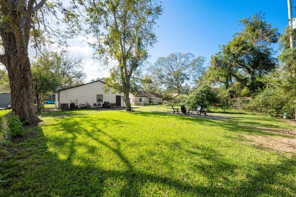 This photo showcases a spacious backyard with lush green grass and mature trees, providing ample shade and a serene atmosphere. The yard includes a cozy seating area with chairs around a fire pit, ideal for outdoor gatherings. The house in the background has a simple, charming design.