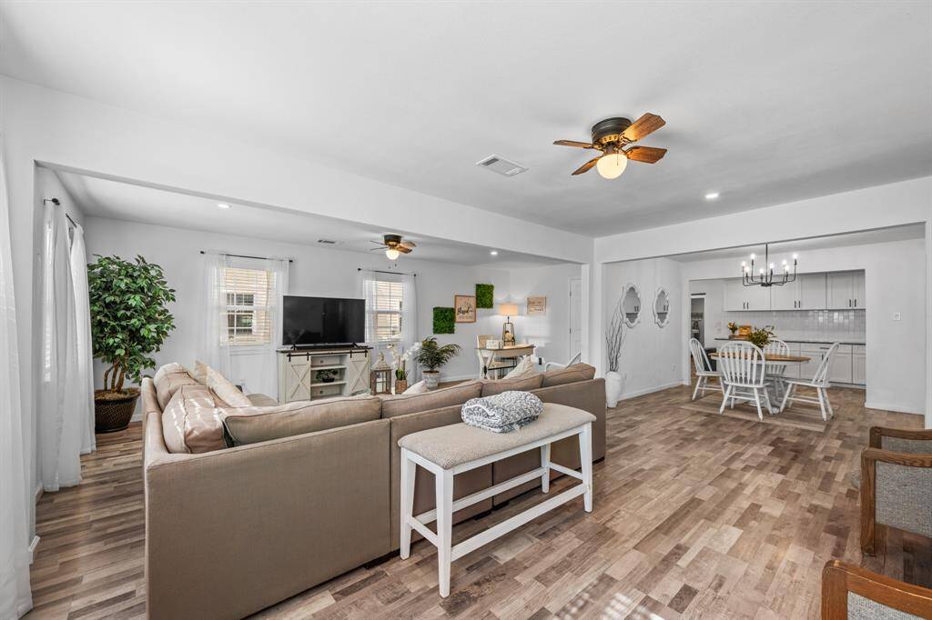 This photo showcases a spacious, open-concept living and dining area with light wood-look vinyl plank flooring. The living room features a large sectional sofa, a TV, and ample natural light from multiple windows. The dining area includes a modern chandelier and a cozy dining set, with a view into the kitchen. The space is bright and inviting, perfect for entertaining.