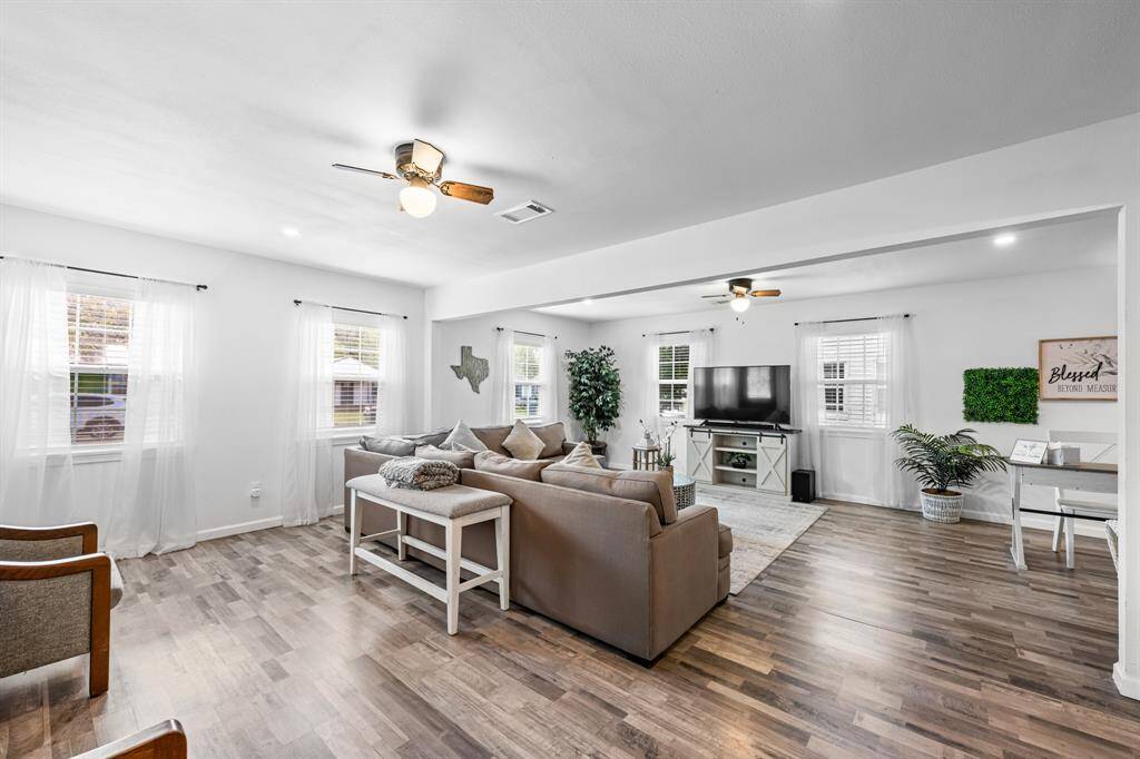 Bright and spacious living room with modern wood-look flooring, neutral tones, and ample natural light. Features ceiling fans, cozy seating area, and a TV setup, creating a welcoming space for relaxation.
