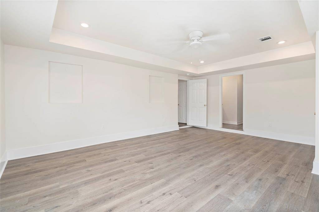 Bright, spacious room with light wood flooring, recessed ceiling lights, and a ceiling fan. The neutral walls and open doorway create a modern, versatile space perfect for various uses.