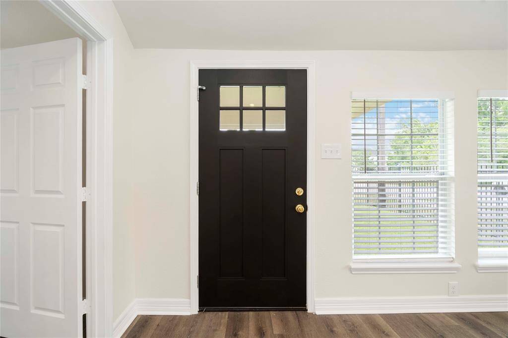 This entryway features a modern black front door with glass panels, allowing natural light to filter in. The space is complemented by light-colored walls, wood flooring, and adjacent windows with blinds, creating a welcoming atmosphere.