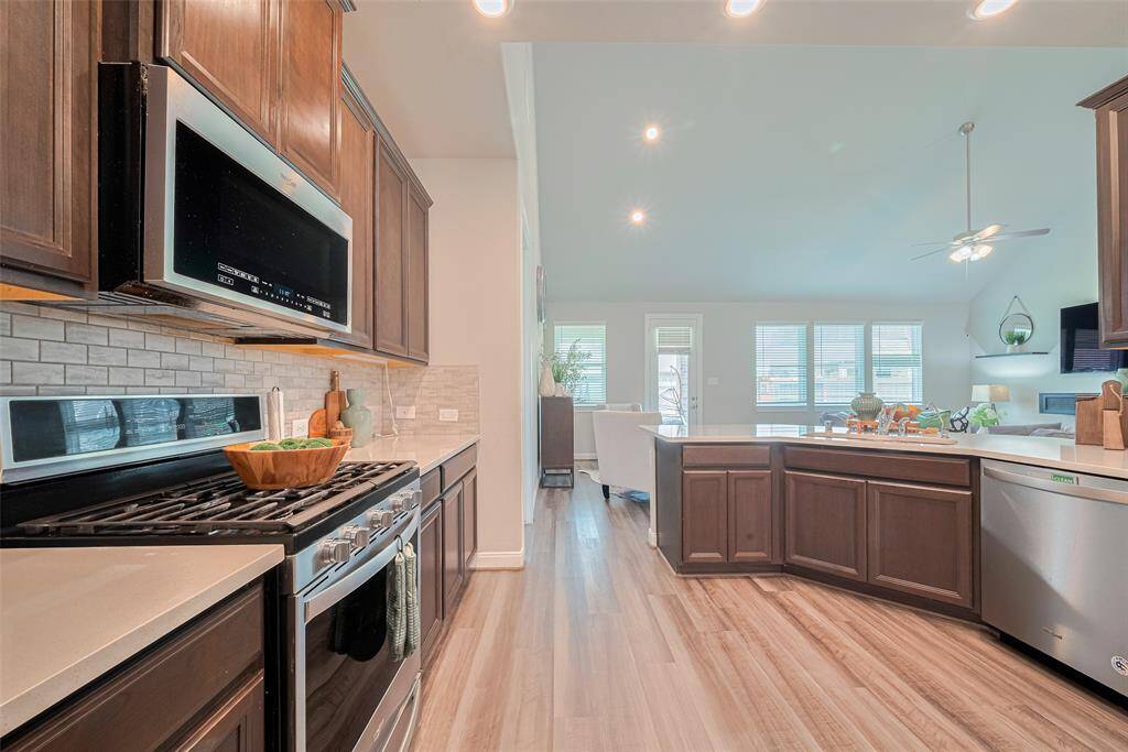 This modern kitchen features sleek dark cabinets, stainless steel appliances, and a spacious open layout leading into a bright living area with large windows and high ceilings. Perfect for entertaining.