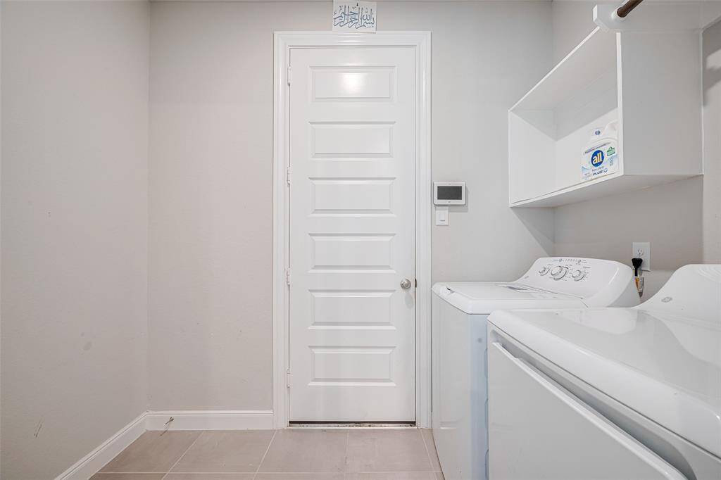 This photo shows a clean, modern laundry room with a washer and dryer set, white shelving for storage. The space is functional with tile flooring and a door leading to the garage.