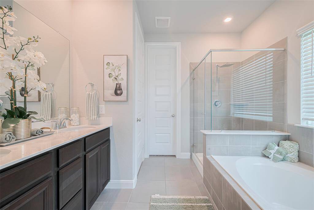 This bathroom features a modern design with a spacious glass-enclosed shower, a separate bathtub, and a dual-sink vanity with dark cabinetry. It has neutral tile flooring, a large mirror, and ample natural light from the window with blinds.