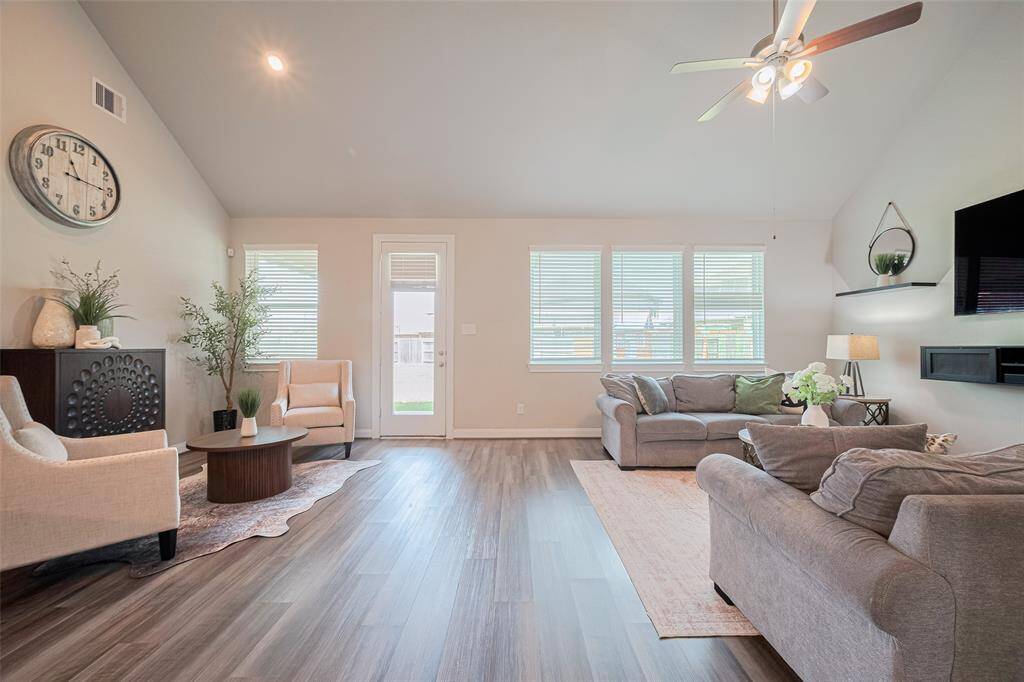 Bright and spacious living room with high ceilings. Features include wood-style flooring, a ceiling fan, and large windows that let in natural light. The room opens to an outdoor space through a glass door.