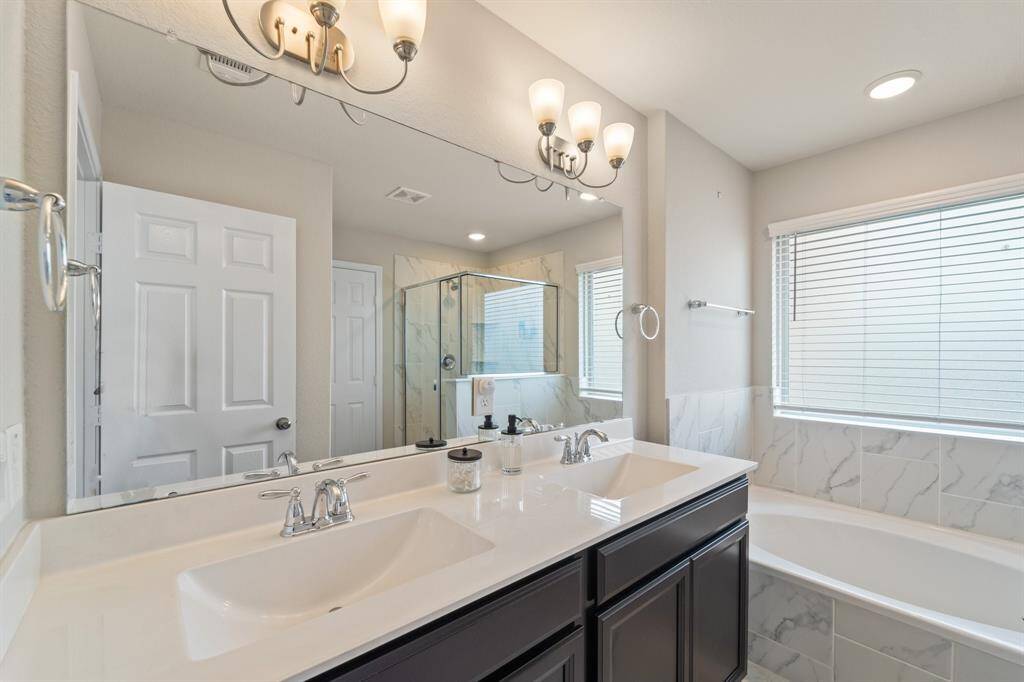 Dual-sink vanity with upgraded lighting sits across from a soaking tub wrapped in marble-look tile—refined and serene.