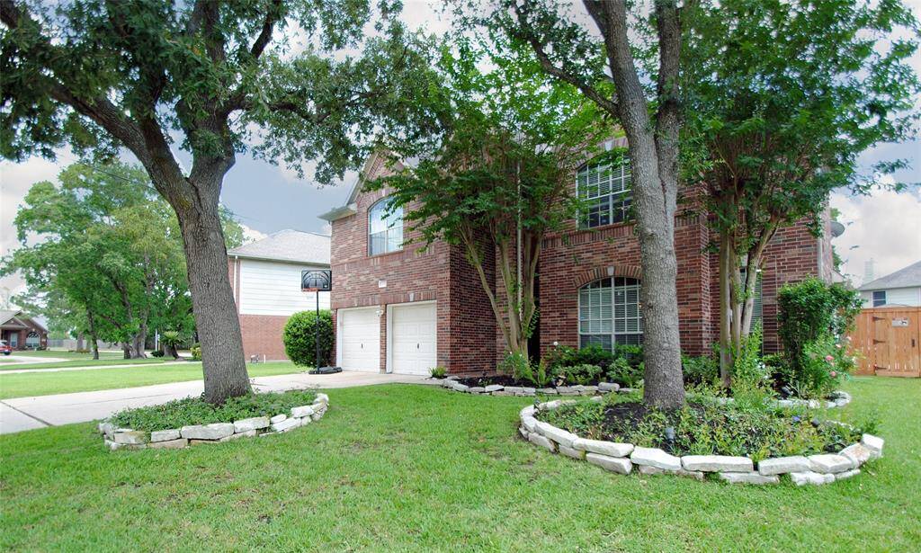 A charming two-story brick home with a well-maintained front yard featuring mature trees and landscaped garden beds. The property includes a two-car garage and a welcoming driveway, creating an inviting curb appeal in a suburban neighborhood.