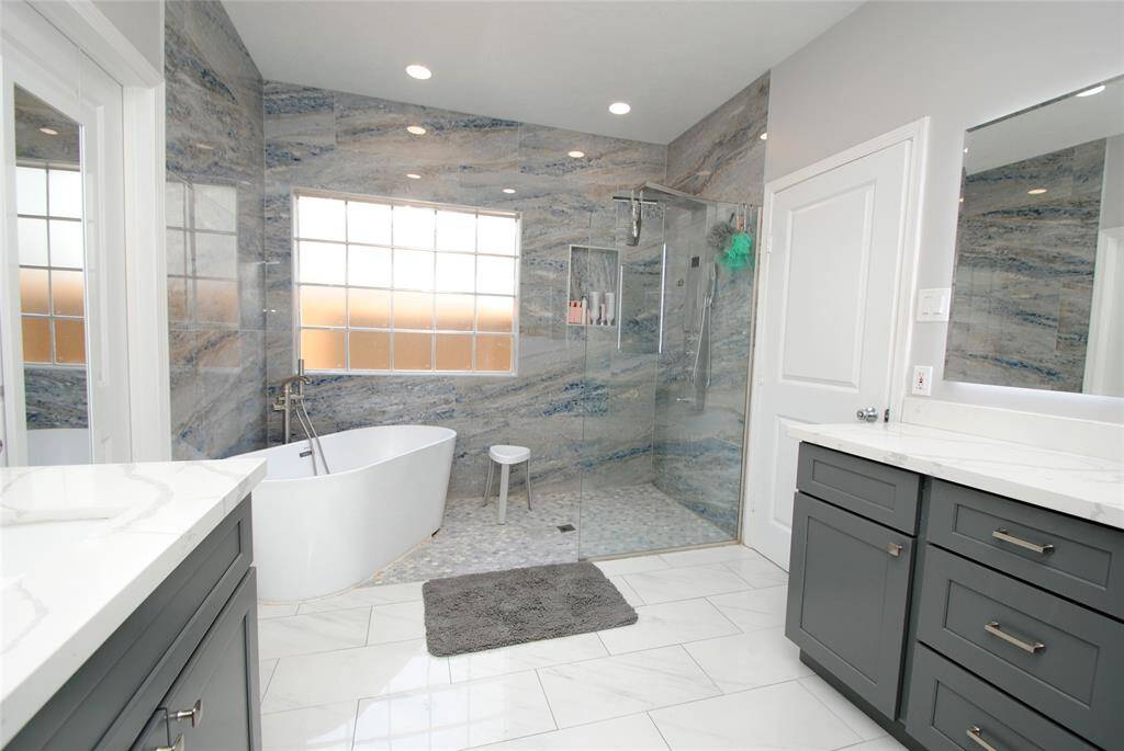This is a modern bathroom featuring dual sinks with sleek gray cabinetry and white countertops. It has a spacious layout with a door leading to the toilet area, bright natural lighting, and a clean, contemporary design.
