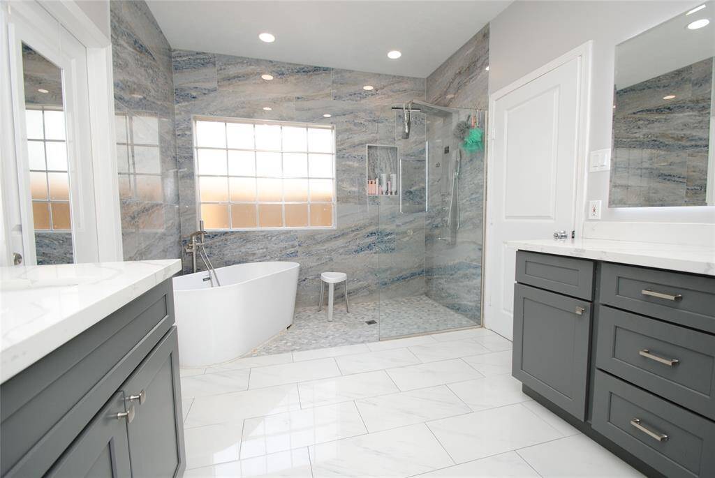 This modern bathroom features a sleek freestanding tub, a spacious glass-enclosed shower, and elegant marble-like wall tiles. It includes dual vanities with ample storage and a bright, airy feel thanks to the large frosted window.