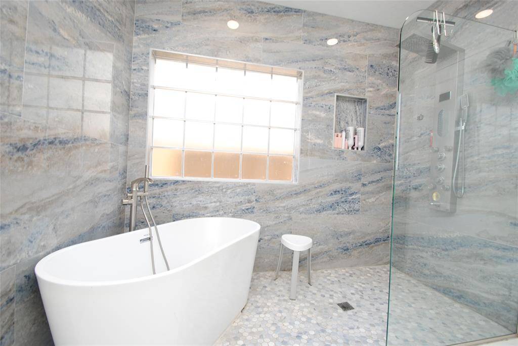 This modern bathroom features elegant marble walls and floors, a sleek freestanding bathtub, and a spacious glass-enclosed shower. Dual vanities with ample storage and a large window provide natural light, creating a luxurious and serene atmosphere.