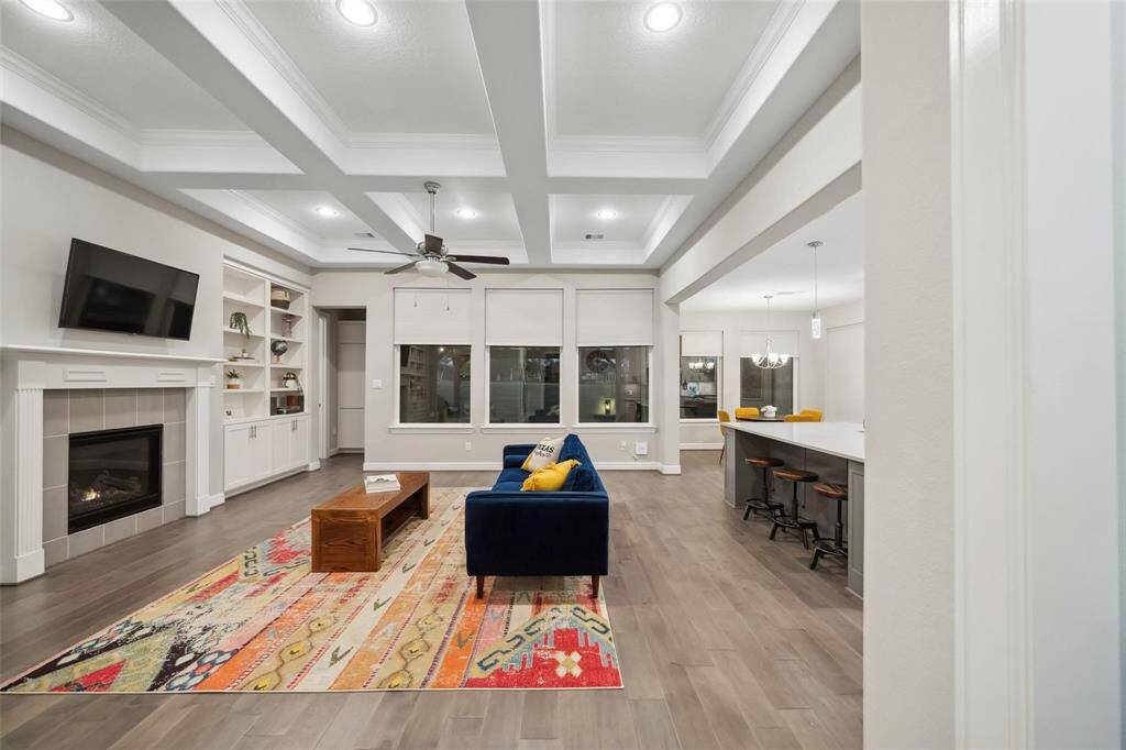 The spacious living room features a beautiful coffered ceiling, a cozy fireplace with a timeless mantle, custom built-ins, and large windows that flood the space with natural light.