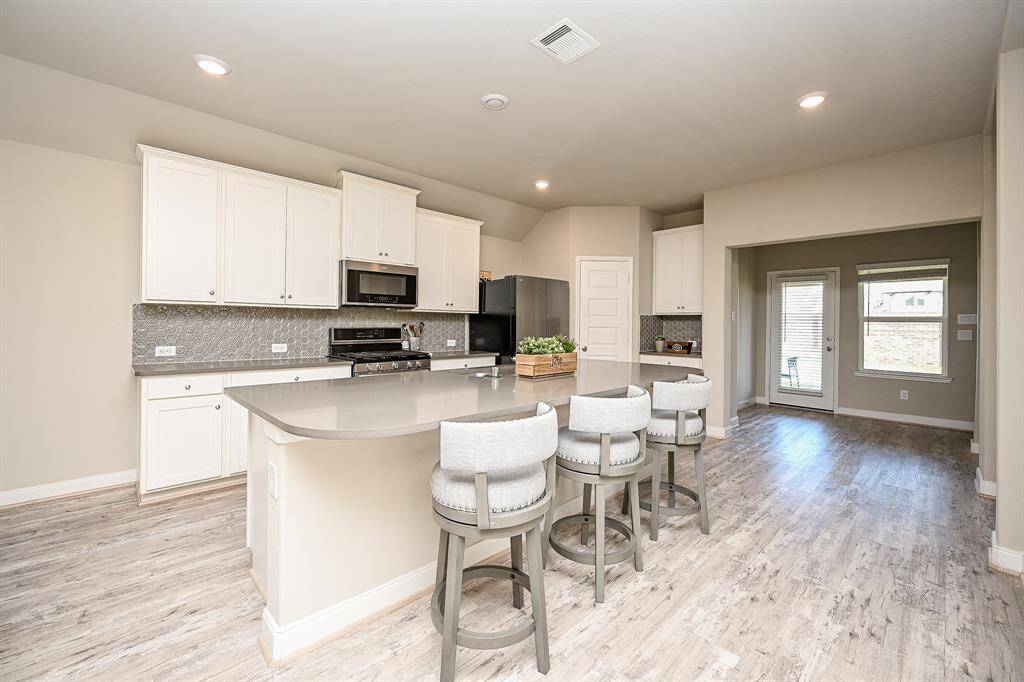 This spacious kitchen features modern white cabinetry, a large island with seating, stainless steel appliances, and stylish backsplash. The open layout flows into a bright dining area with access to the backyard through a glass door.