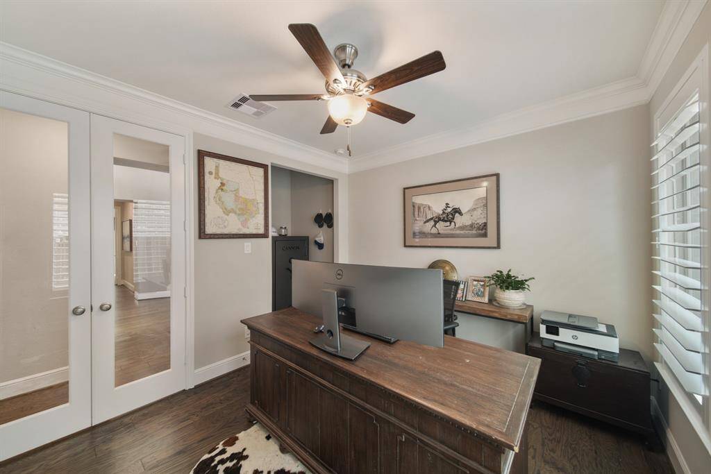 The sophisticated home office is set behind elegant double glass doors, featuring classic crown molding, rich hardwood floors, and custom shutters that offer both style and privacy—perfect for work or quiet reflection.