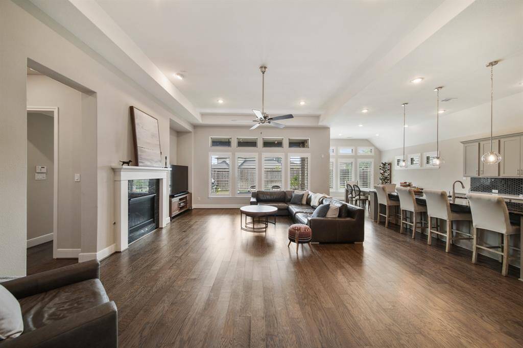 Step into the expansive open-concept living, breakfast, and kitchen area, where soaring ceilings and a wall of windows at the rear flood the space with natural light. A stylish fireplace anchors the living area, while rich wood floors flow seamlessly throughout, creating a warm and inviting ambiance.